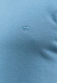 Jasnoniebieska, dziana koszula z fakturowanej tkaniny. Posiada małe, wyszywane logo konia na klatce piersiowej.