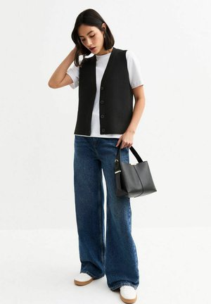 Zwarte mouwloze vest met knopen, gecombineerd met een wit T-shirt, wijde blauwe jeans en een kleine zwarte handtas. Textuur van de materialen zichtbaar.