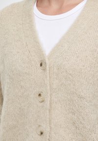 Gros plan d'un cardigan beige en maille duveteuse avec trois boutons ronds porté sur un t-shirt blanc côtelé à col rond.