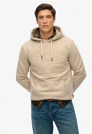 Beige hoodie met reliëf merklogo, verstelbare koordjes en voorzakken, gedragen over een geruite shirt en blauwe spijkerbroek.