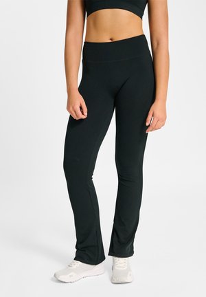 Frau trägt schwarze, hoch taillierte, ausgestellte Trainingsleggings und weiße Sportschuhe, steht vor einem schlichten hellen Hintergrund.