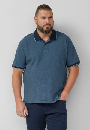 Brodaty mężczyzna w niebieskiej, paskowanej koszulce polo z krótkim rękawem i ciemnych spodniach, stojący z jedną ręką w kieszeni na jednolitym tle.