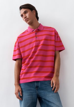 Ung mand iført lyserød og rødrandet polo og blå jeans, stående mod en hvid baggrund og kigger til siden.