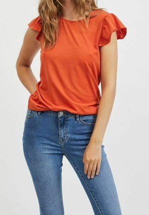 Kobieta ubrana w pomarańczową bluzkę z krótkim rękawem z falbanami oraz niebieskie dżinsy, stoi z jedną ręką w kieszeni, a drugą opartą luźno wzdłuż ciała.