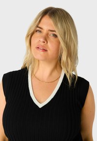 Černý, žebrovaný top bez rukávů s bílým V-krčem. Modelka nosí tenký zlatý řetízek. Světle vlnité blond vlasy.