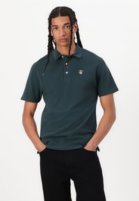 Mørkegrønn polo skjorte laget av bomull med en tre-knapps placket. Har en liten logo på venstre bryst og korte ermer.