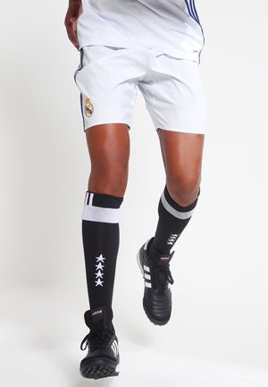 Jambes d'une personne portant un short blanc du Real Madrid, des chaussettes de football noires avec des étoiles blanches et des chaussures de football Adidas noires.