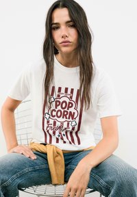 T-shirt en coton blanc avec un graphique de popcorn rouge et blanc et le texte "Crispy Dreams Popcorn Fresh & Free". Le modèle porte un jean bleu et un pull beige.
