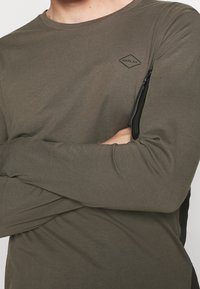 Olivegrünes Longsleeve-Shirt mit rundem Ausschnitt. Verfügt über eine schwarze Reißverschlusstasche am linken Arm und ein kleines Diamant-Logo auf der Brust.