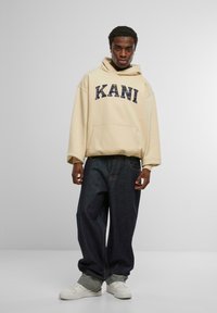 Beige hoodie med "KANI" tryckt i en mörk färg, med en framficka och lös passform, kombinerad med mörkblå jeans och vita sneakers.
