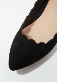 Zapato plano negro de ante con punta afilada y detalle de borde con forma de ondas, y forro interior beige.