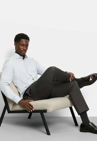 Hellblau gemustertes Hemd mit Knöpfen, dunkelbraune Hosen und polierte Schuhe. Das Modell sitzt entspannt auf einem minimalistisch gestalteten Stuhl mit cremefarbenem Kissen.