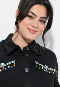 Mustat farkkutakki, jossa on rispaantunut kaulus, koristeelliset helmikoristeet taskuissa ja hopeansävyiset kiinnitykset.