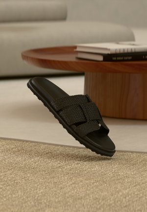 Fekete szövött bőr papucs lebeg bézs szőnyeg felett, háttérben egy kerek fából készült dohányzóasztallal és egymásra rakott könyvekkel, homályos fókuszban.