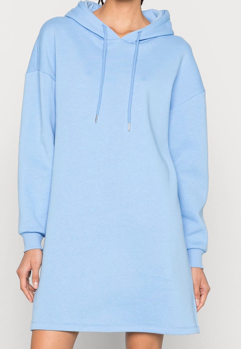Femme portant une robe sweat à capuche oversize bleu clair avec manches longues et cordons à l'avant.