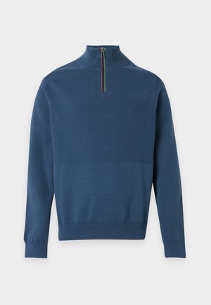 Homme debout, mains dans les poches, portant un pull bleu à fermeture éclair quart, pantalon noir et mocassins jaunes sur fond blanc.