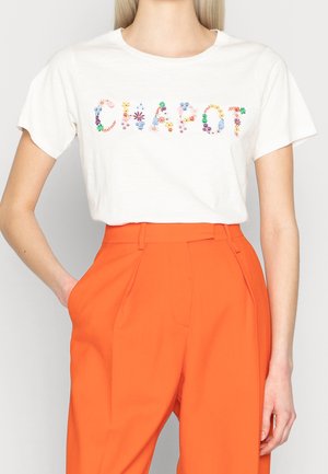 Femme portant un T-shirt blanc avec "CLAROT" écrit en lettres florales colorées et un pantalon taille haute orange vif, une main dans la poche.