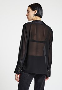 Blusa nera trasparente con maniche lunghe, polsini con doppio bottone e scollo con colletto. Il tessuto traslucido lascia intravedere un reggiseno nero sottostante.
