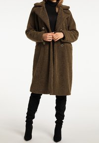 Brun, struktureret lang frakke med stor krave, dobbelte knapper og bælte. Båret over en sort rullekrave, parret med sorte knæhøje støvler.