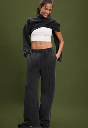 Lächende Frau in schwarzen weiten Hosen, weißem Cropped-Top und schwarzem Sweatshirt, das über den Schultern hängt, vor olivfarbenem Hintergrund.