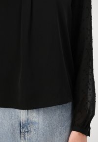 Svart topp med en glatt kropp og gjennomsiktige, utsmykkede ermer som har små perler og en samlet mansjett. Kombinert med lyseblå denim.