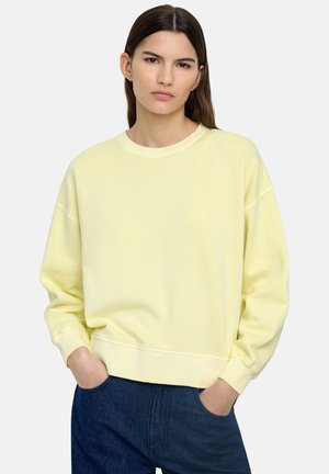 Młoda kobieta z długimi brązowymi włosami, ubrana w jasnożółtą bluzę i ciemnoniebieskie dżinsy, ręce w kieszeniach, neutralne tło.