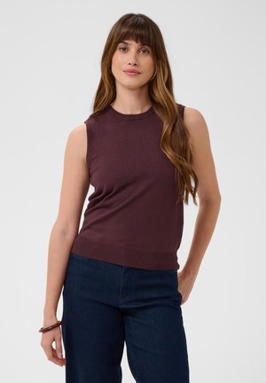 Femme aux longs cheveux bruns portant un haut sans manches violet foncé et un pantalon bleu foncé, debout devant un fond clair uni.