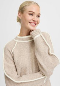 Beige gebreide trui met witte accenten, voorzien van een ronde halslijn, raglanmouwen en geribbelde boorden. Zachte textuur en een comfortabele pasvorm.
