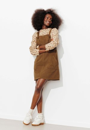Femme avec une coiffure afro portant un chemisier à fleurs, une robe marron sans manches, des chaussettes blanches et des baskets, appuyée contre un mur blanc, souriante.
