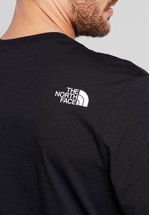 Mand iført sort t-shirt med hvidt The North Face-logo på øverste del af ryggen, fotograferet bagfra og fra siden.