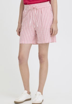 Person iført rød-hvide, stribede shorts med snor, rød top og hvide sneakers mod en ensfarvet baggrund.