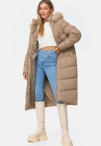 Beige gepolsterter Mantel mit einer Kunstfellkapuze, mit Reißverschluss vorne und seitlichen Taschen, getragen über blauen hoch taillierten Jeans und cremefarbenen Plateaustiefeln.