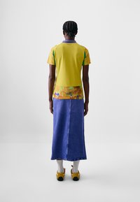 Camicetta gialla a maniche corte con accenti verdi e un motivo colorato sul fondo, abbinata a una lunga gonna di jeans blu e scarpe gialle.