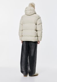 Puffermantel in hellem Beige mit Kapuze, ausgestattet mit gesteppten Abschnitten. Kombiniert mit weiten grauen Jeans und hellen Schuhen. Minimalistisches Design.