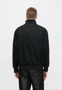 Μαύρο μπουφάν bomber με ψηλό γιακά, ριμπ μανσέτες και τελείωμα. Διαθέτει μινιμαλιστικό σχεδιασμό και λεπτομέρεια λογότυπου στο λαιμό. Φορεμένο με σκουρόχρωμο, λαμπερό παντελόνι.