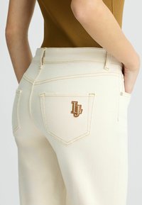 Jean crème avec une coupe ajustée, des coutures dorées fines et un accent de logo doré sur la poche arrière. Texture lisse.