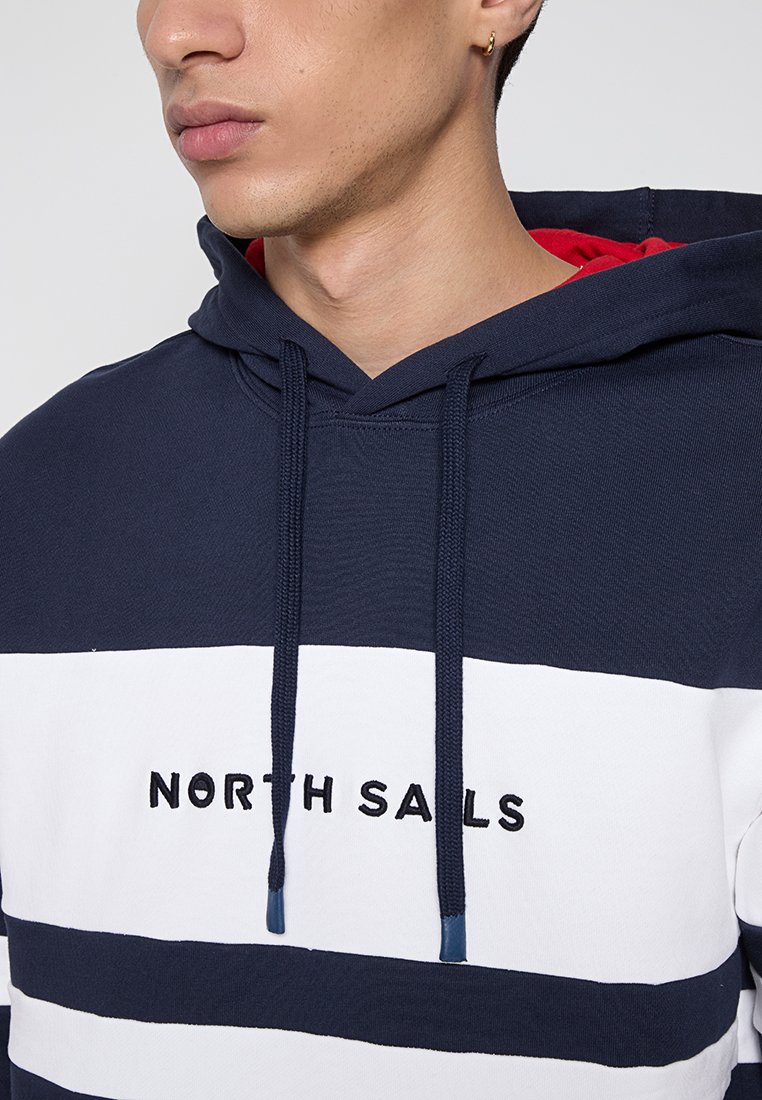 Giovane che indossa un felpa a righe blu navy e bianche con la scritta "North Sails" sul petto e l'interno del cappuccio rosso, rivolto a sinistra.