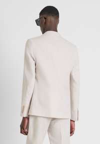 Veste de costume en lin beige clair avec une coupe ajustée, fermeture à un bouton, revers crantés et détails de couture visibles sur les manches.