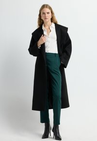 Zwart wollen jasje met brede revers, gecombineerd met een witte knopshirt en teal tailored broek. Zwarte stilettohakken maken de outfit compleet.