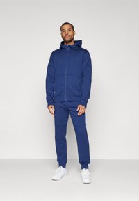 Mörkblå huvtröja med dragkedja och matchande joggers, tillverkad av mjukt material. Har ribbade ärmslut och en ficka på vänster sida.