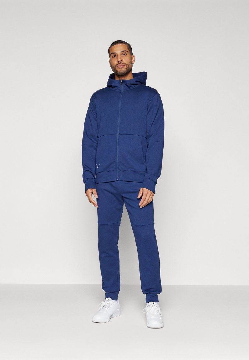 Mörkblå huvtröja med dragkedja och matchande joggers, tillverkad av mjukt material. Har ribbade ärmslut och en ficka på vänster sida.