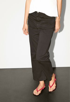 Schwarze weite Baumwollhosen mit Knopfverschluss und klassischem Fünf-Taschen-Design, kombiniert mit rosa Zehentrenner-Sandalen.
