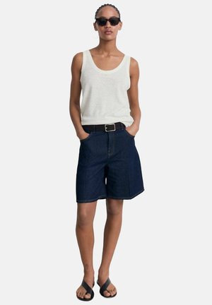 Top senza maniche grigio chiaro, pantaloni shorts in denim blu scuro, cintura nera e occhiali da sole neri. Modella in piedi con le mani nelle tasche, indossando sandali piatti.