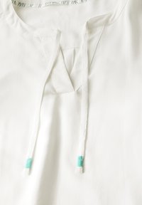 Witte tuniektop met een V-halslijn, voorzien van een voorkeursplit, striksluiting met turquoise accenten en een zachte stofstructuur.