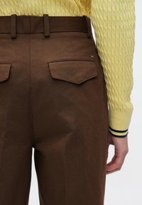 Καφέ παντελόνι με λείο, δομημένο ύφασμα, που διαθέτει δύο πίσω τσέπες και διακριτική μεταλλική λεπτομέρεια, σε συνδυασμό με κίτρινο πουλόβερ με υφή.