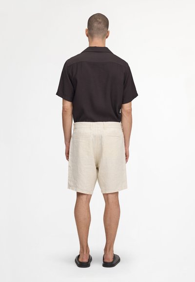 Homme debout, dos tourné vers la caméra, portant une chemise noire à manches courtes, un short beige et des chaussures noires sans lacets sur un fond blanc uni.