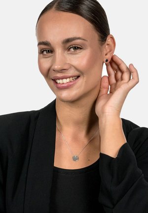 Silberner Anhänger-Halsreif an einer zarten Kette, getragen mit einem schwarzen Oberteil und Blazer. Schwarze Steckohrringe. Glatte Hautfarbe. Minimalistisches Design.