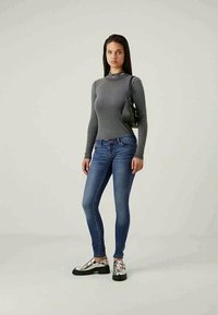Top de manga larga gris con relieve, combinado con jeans ajustados azules y zapatos metálicos con tiras. La modelo sostiene un pequeño bolso negro.