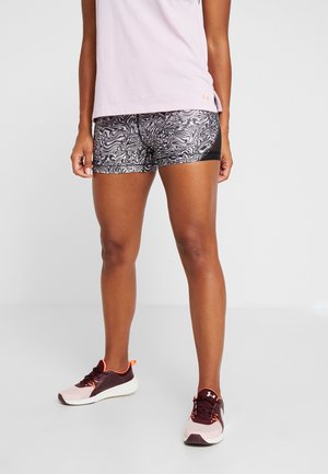 Person iført sorte og hvide mønstrede atletikshorts, lys rosa top og mørk rosa og hvide løbesko, står på en lys gulv.