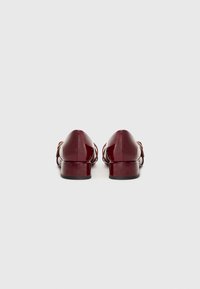 Zapatos de charol burdeos con un tacón bajo. Presentan tiras cruzadas y una punta redondeada, ofreciendo una silueta elegante.